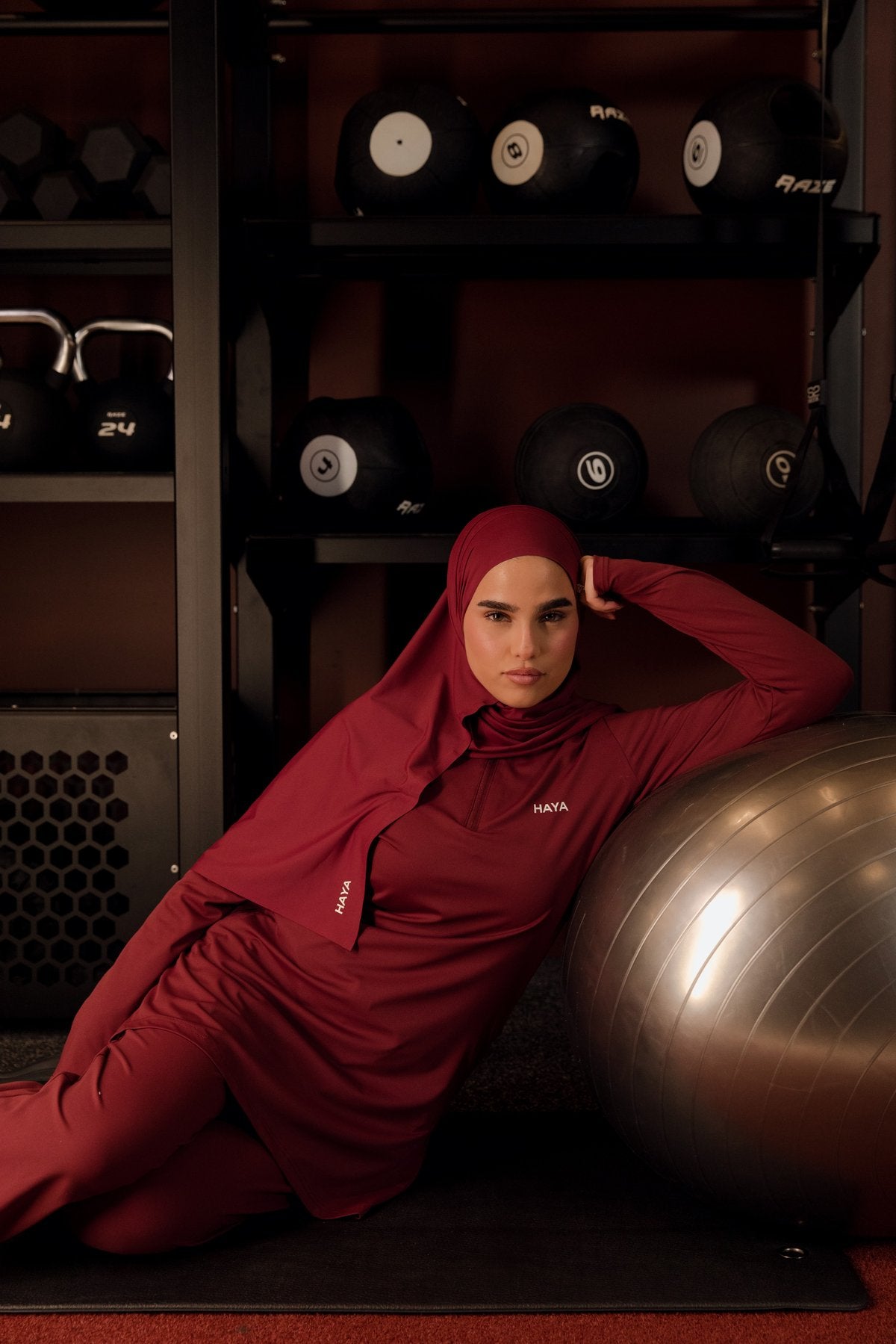 A woman in a red athletic hijab and top rests against a silver exercise ball in a gym.